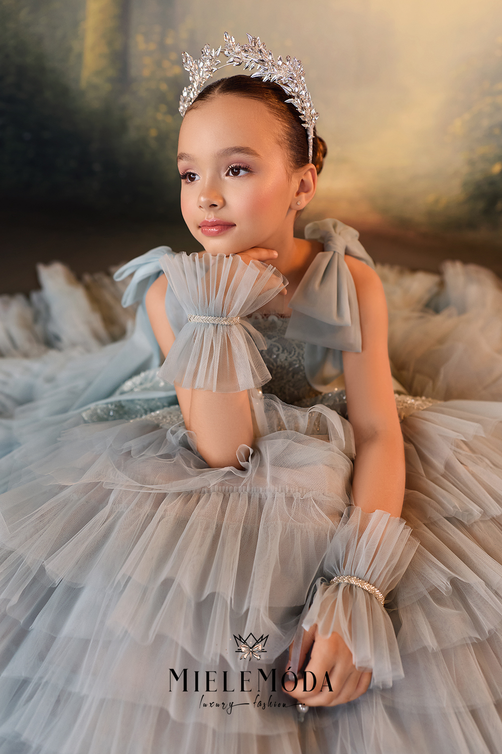 A young girl wearing a sparkling silver tiara and a voluminous light blue-gray tulle ball gown with matching ruffled gloves, posed against a soft, painterly forest background.