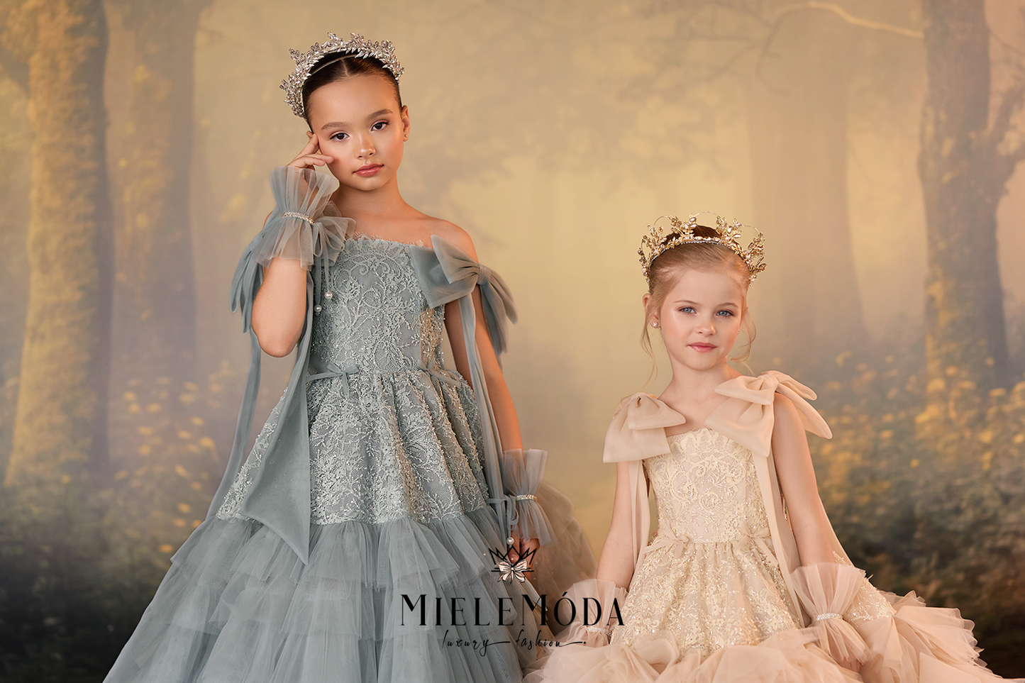 Two young girls wearing elegant fantasy princess dresses and tiaras, posed against a soft, misty forest backdrop. The girl on the left wears a dusty blue gown with sheer cape sleeves, while the girl on the right wears a champagne beige gown with similar sheer sleeves.