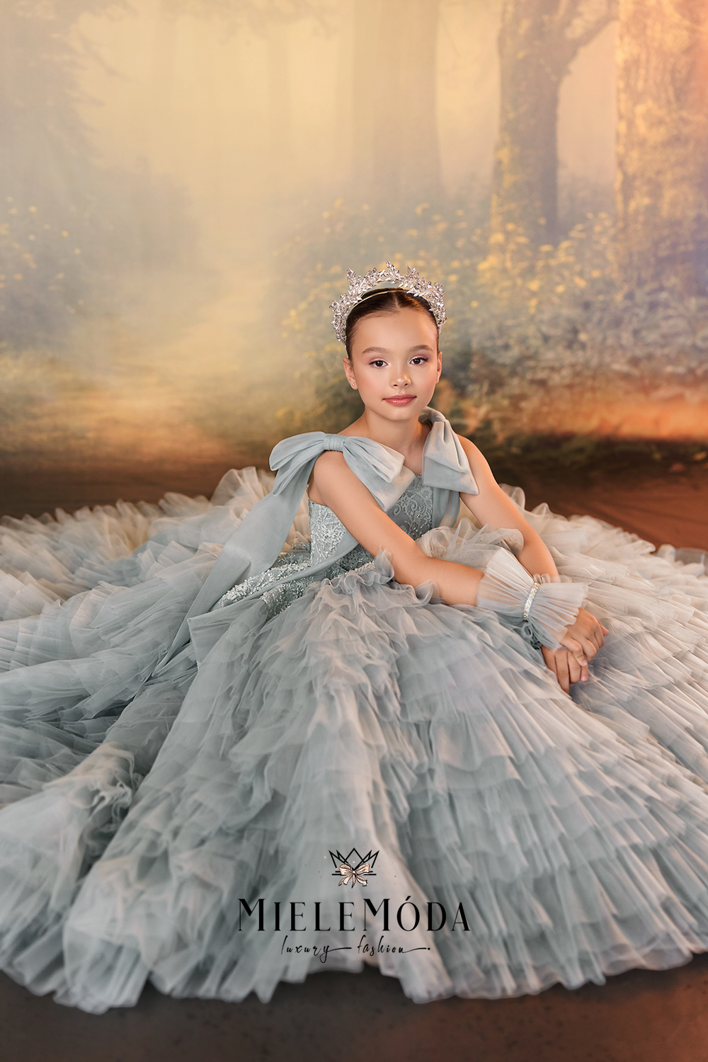 A young girl wearing a voluminous light blue tulle ball gown with a tiara and white gloves, posed in front of a warm-toned forest backdrop. The brand name MIELEMODA is visible at the bottom.