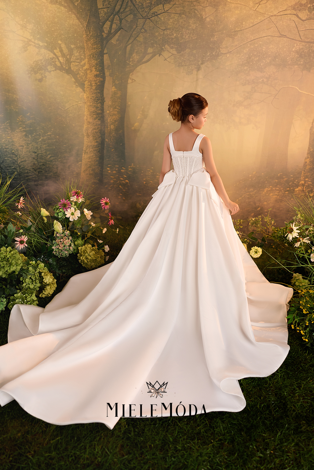 A woman wearing a white sleeveless wedding dress with a long train stands in a misty forest setting with flowers and trees in the background. The brand name MIELEMÓDA is printed on the dress.