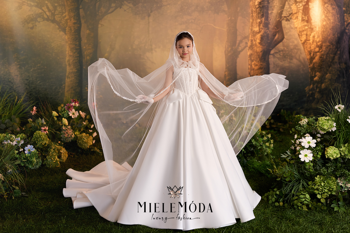 A young girl wearing a white ballgown wedding dress with a sheer veil, standing in a forest-themed studio setting with flowers and dramatic golden lighting.