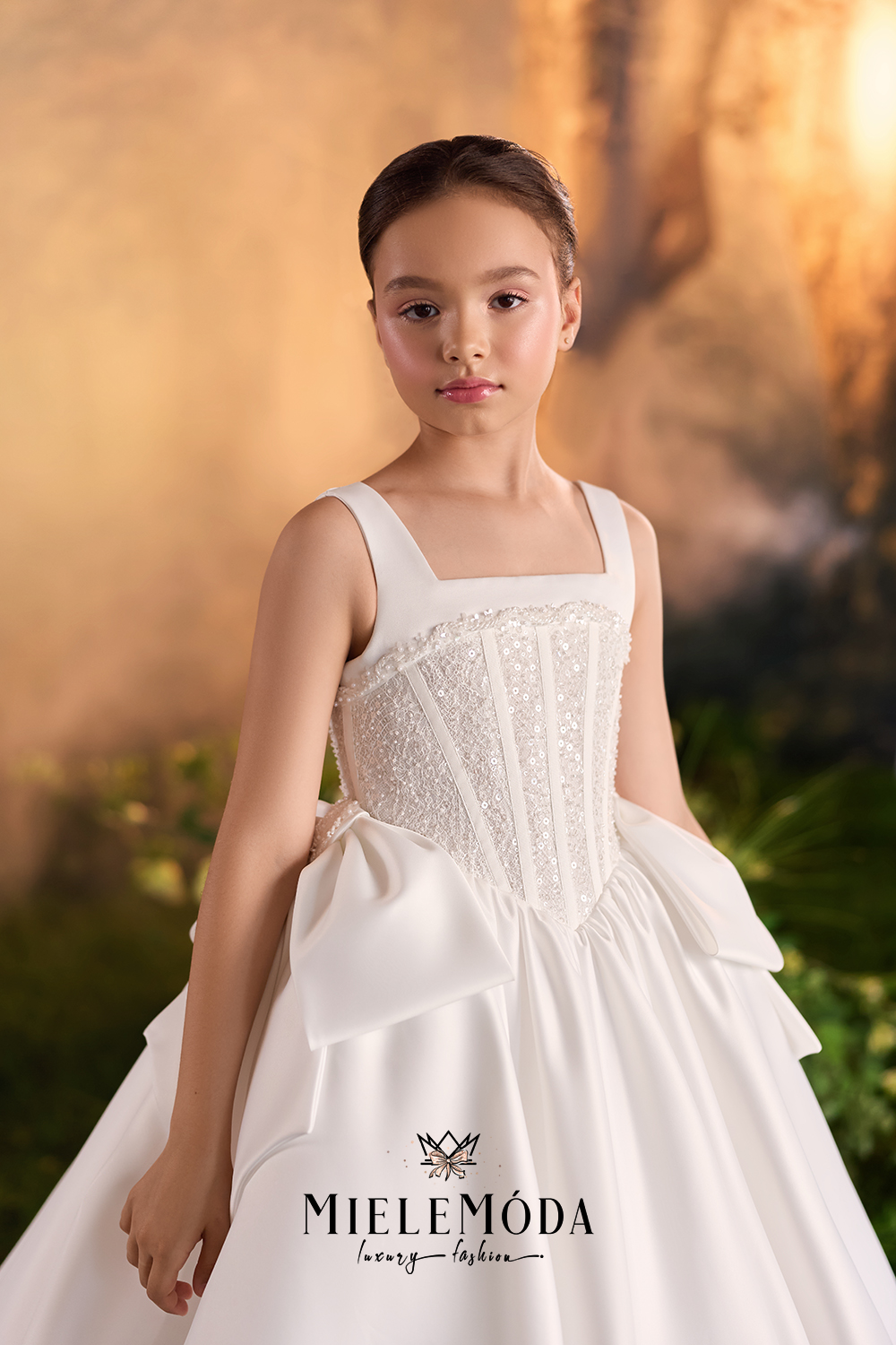 A young girl wearing a white sleeveless ball gown with a lace corset bodice and a full layered skirt, standing against a warm golden outdoor background. The brand name 'MIELEMÓDA' is printed on the skirt.