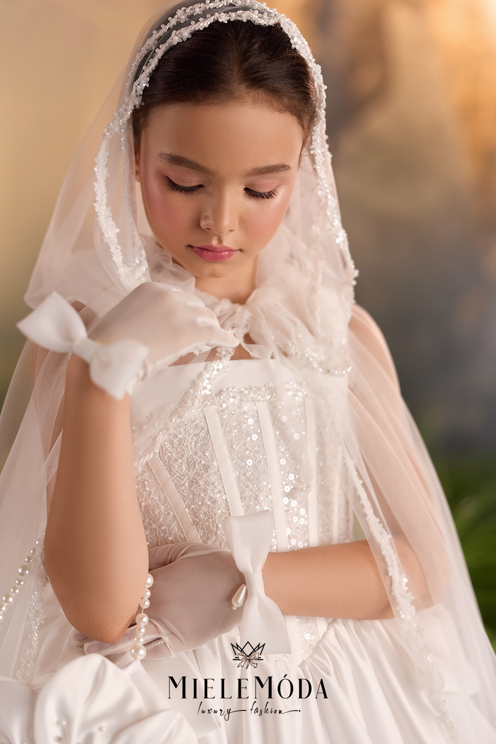 A young girl wearing a white First Communion dress with lace, beadwork, a matching veil, and white gloves, posed against a warm, softly blurred background. The brand name 'MieleMóda' is visible at the bottom.
