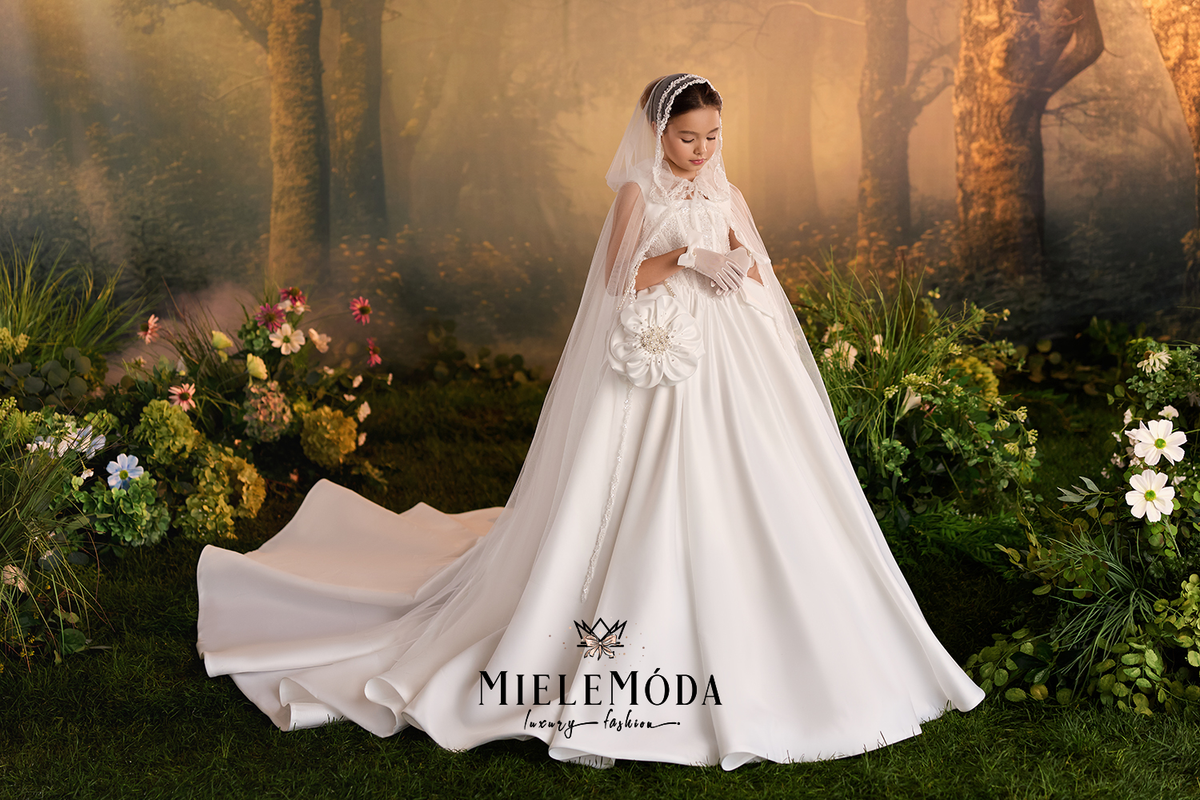 A young girl wearing a white wedding dress with a long train, veil, and flower bouquet stands in a forest-themed setting with flowers and trees. The brand name MIELEMÓDA is printed on the dress.