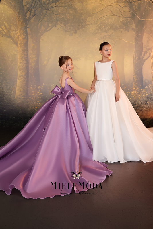 Two young girls modeling formal ball gowns in a studio setting with a forest backdrop. The girl on the left wears a lavender-purple satin gown with a large bow at the back, and the girl on the right wears a sleeveless white satin gown with a fitted waist and full skirt.