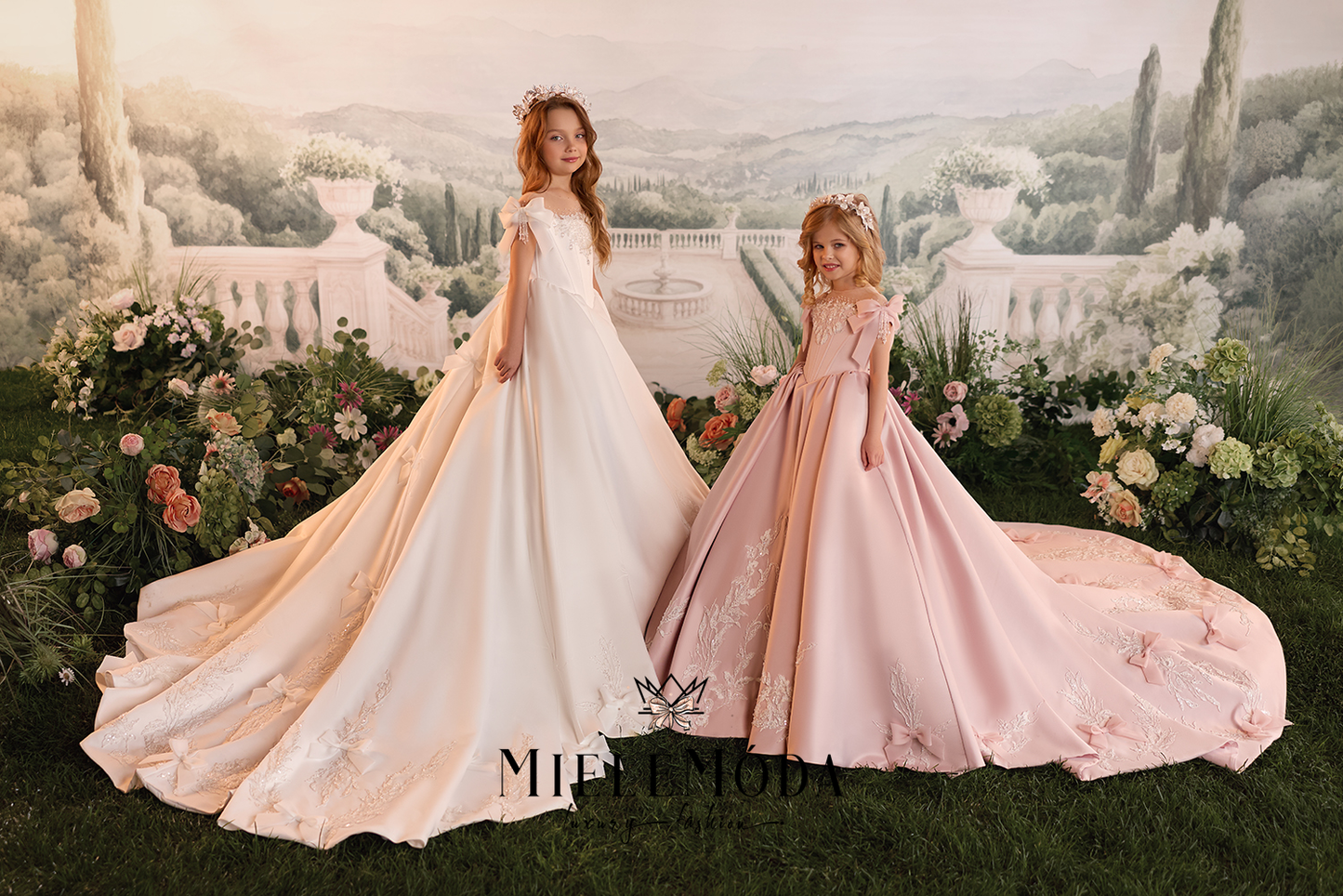 Two young girls wearing elegant ballgown wedding dresses pose in front of a scenic garden backdrop. The girl on the left wears a cream-to-ivory gown with peach undertones, and the girl on the right wears a soft blush pink gown.