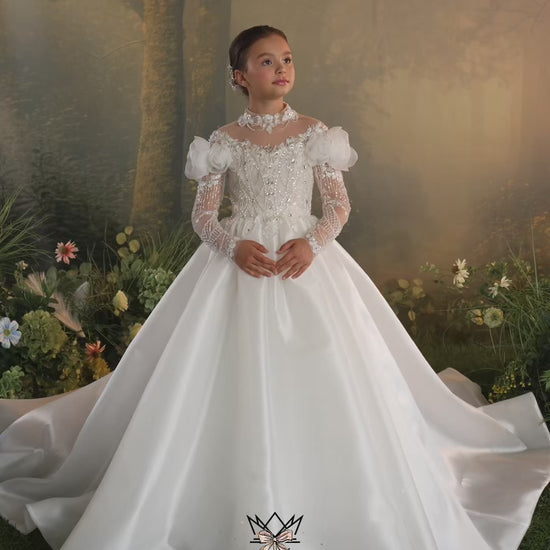 A young girl modeling a white off-the-shoulder ball gown with intricate lace and bead embroidery, set against a soft, warm-toned forest backdrop.
