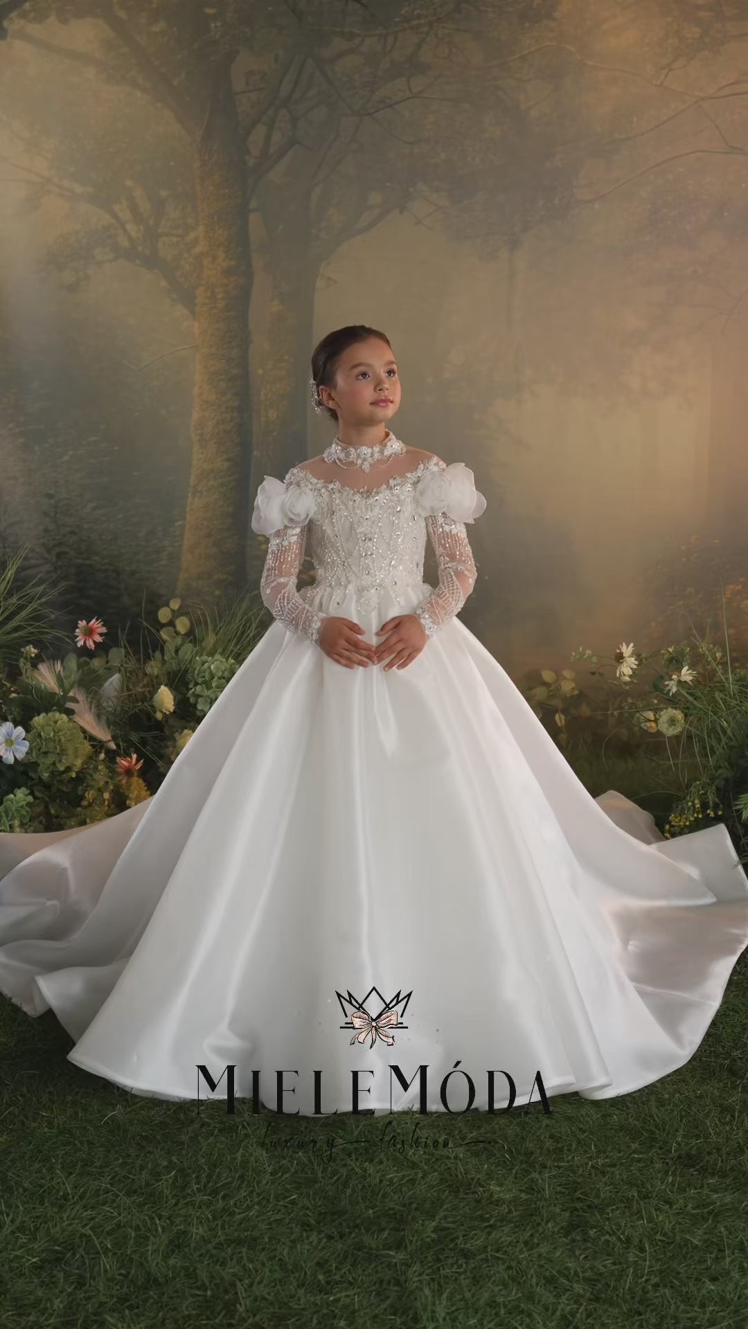 A young girl modeling a white off-the-shoulder ball gown with intricate lace and bead embroidery, set against a soft, warm-toned forest backdrop.