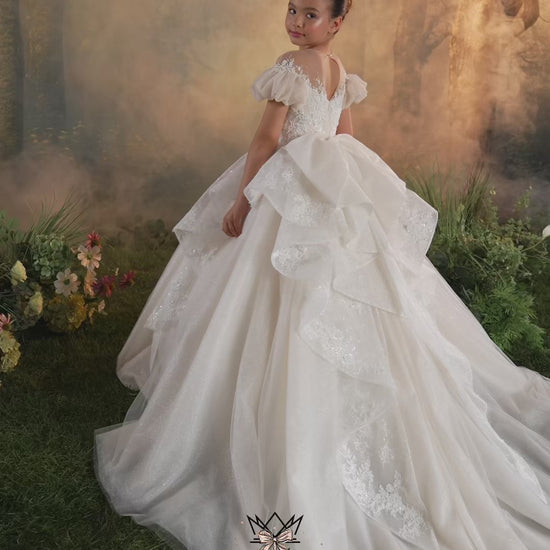 A young girl wearing a formal off-the-shoulder lace ball gown with sheer floral embroidery and a matching veil, posed against a warm golden outdoor backdrop. The brand name MieleModa is visible at the bottom.