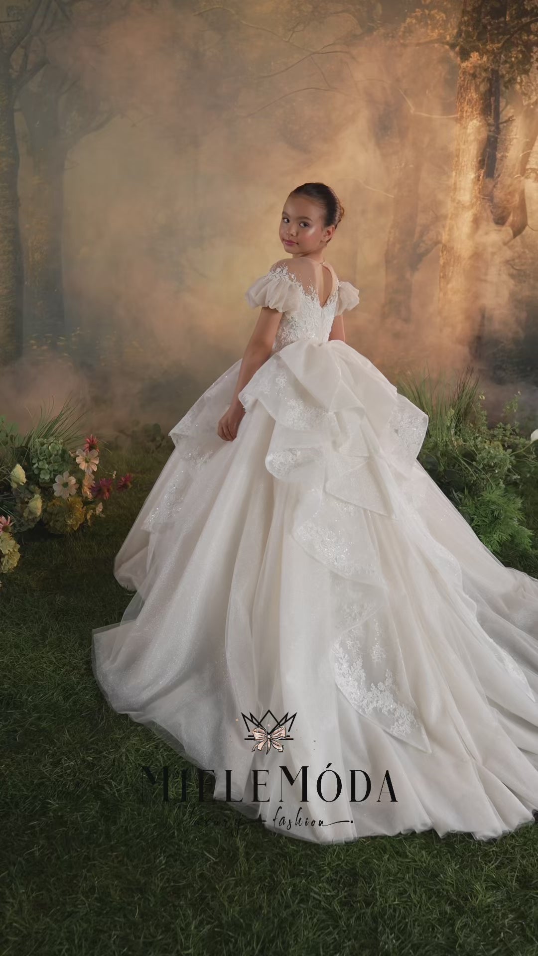 A young girl wearing a formal off-the-shoulder lace ball gown with sheer floral embroidery and a matching veil, posed against a warm golden outdoor backdrop. The brand name MieleModa is visible at the bottom.
