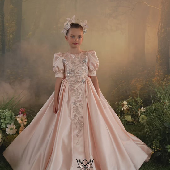 Two young girls modeling a pale pink ball gown with intricate silver and beaded embroidery on the bodice and a matching floral headpiece, set against a soft-focus forest backdrop. The brand name 'MieleMóda' is visible at the bottom.