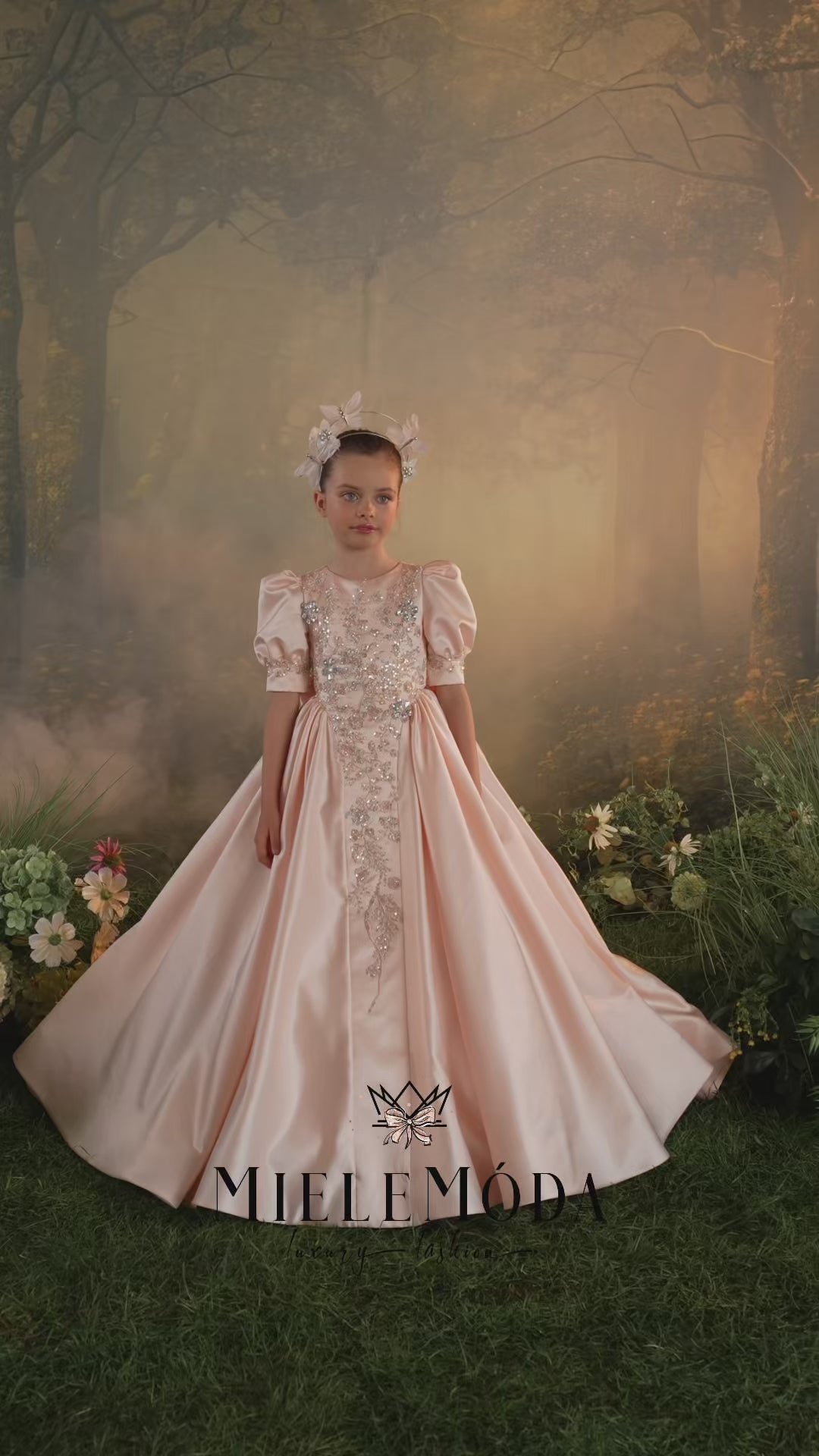 Two young girls modeling a pale pink ball gown with intricate silver and beaded embroidery on the bodice and a matching floral headpiece, set against a soft-focus forest backdrop. The brand name 'MieleMóda' is visible at the bottom.