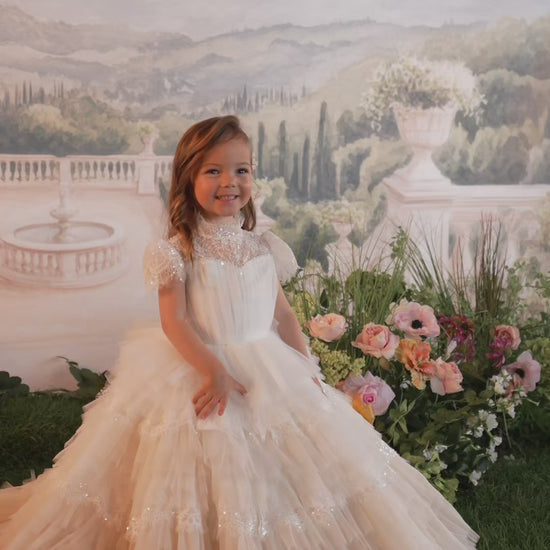 Two young girls wearing elegant white lace and tulle first communion dresses, posed in front of a scenic garden backdrop. The brand name 'MIELEMODA' is displayed at the bottom.