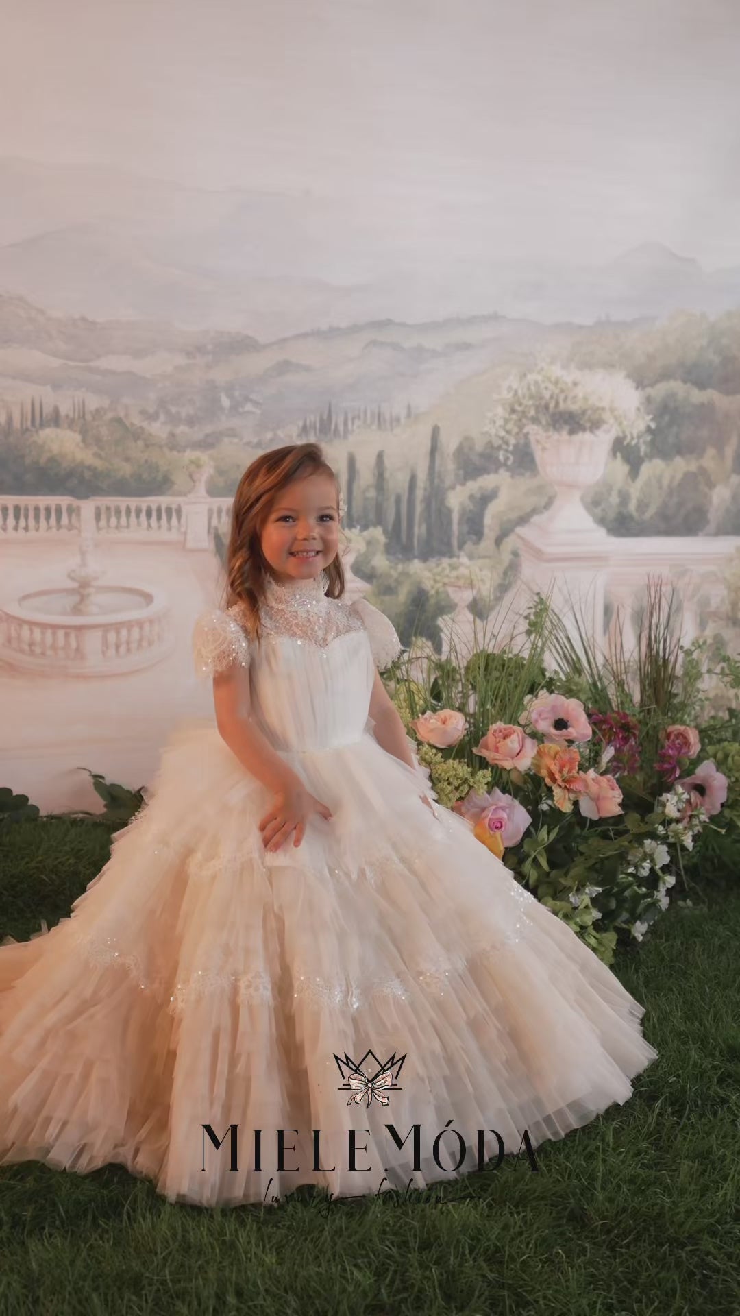 Two young girls wearing elegant white lace and tulle first communion dresses, posed in front of a scenic garden backdrop. The brand name 'MIELEMODA' is displayed at the bottom.