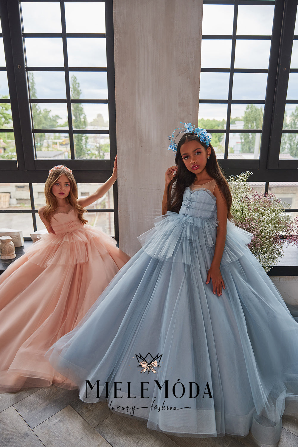 beautiful girls sitting by large window wearing tulle couture dresses in pink and blue modeling for miele moda boutique